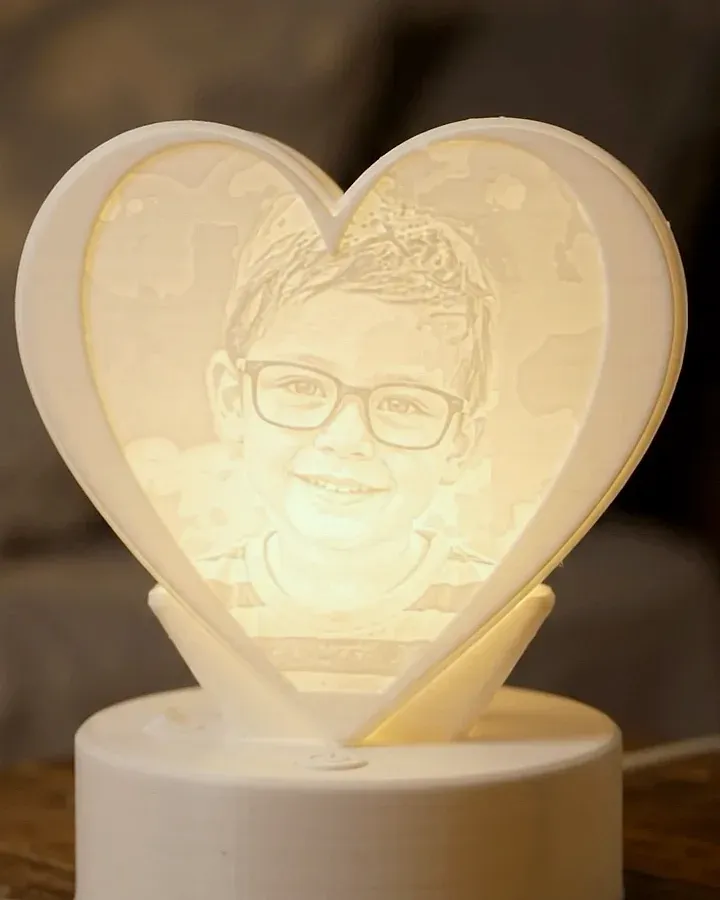 Une lithophanie en forme de cœur allumée sur une table de chevet, la photo d'un enfant à lunettes apparaît dans le plastique blanc, posée sur un socle lumineux rond